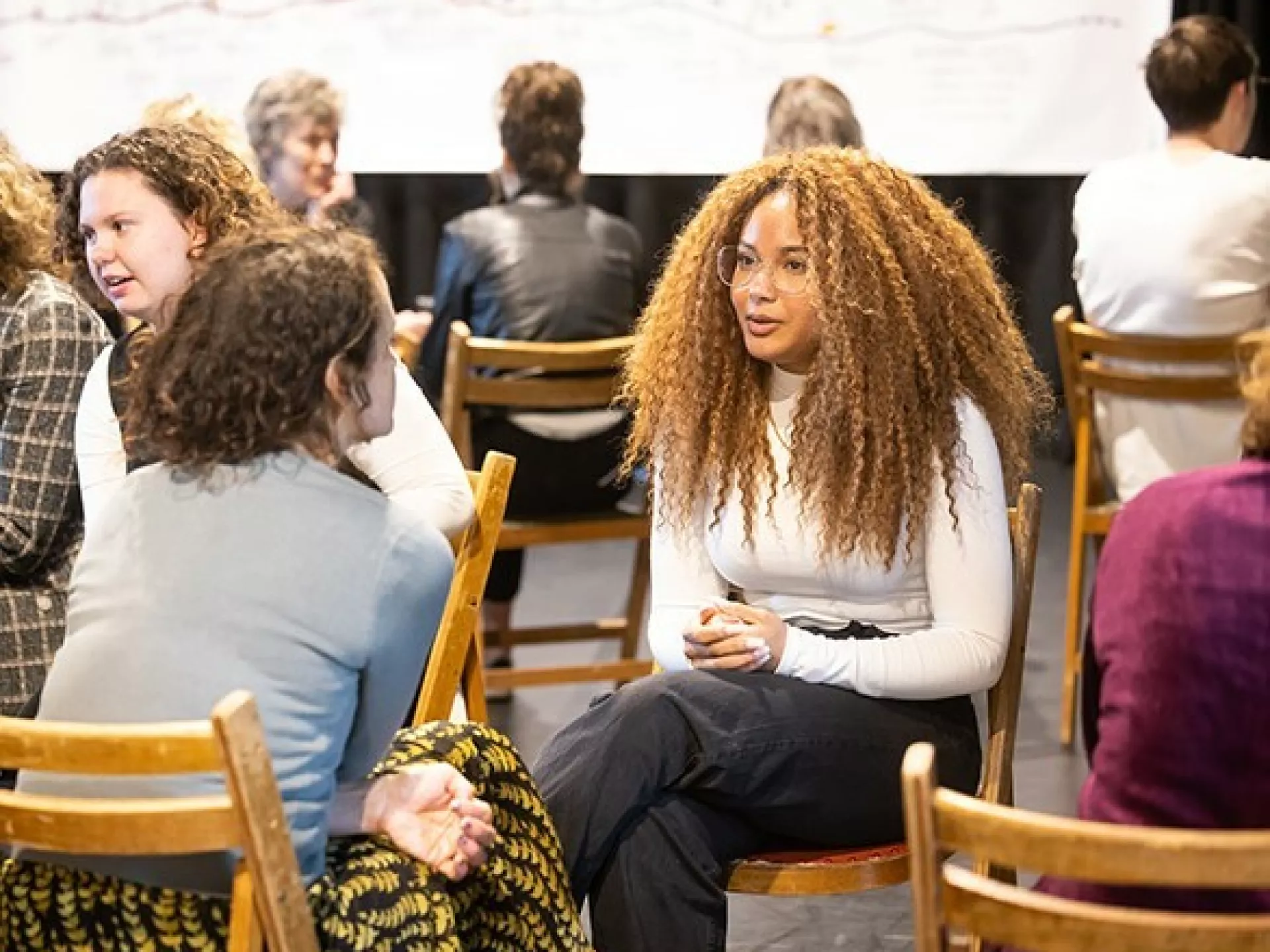 De Onmisbare Schakel - Drenthe - Twee mensen praten met elkaar. Ze zitten in een zaal waar mensen een workshop aan het volgen zijn.