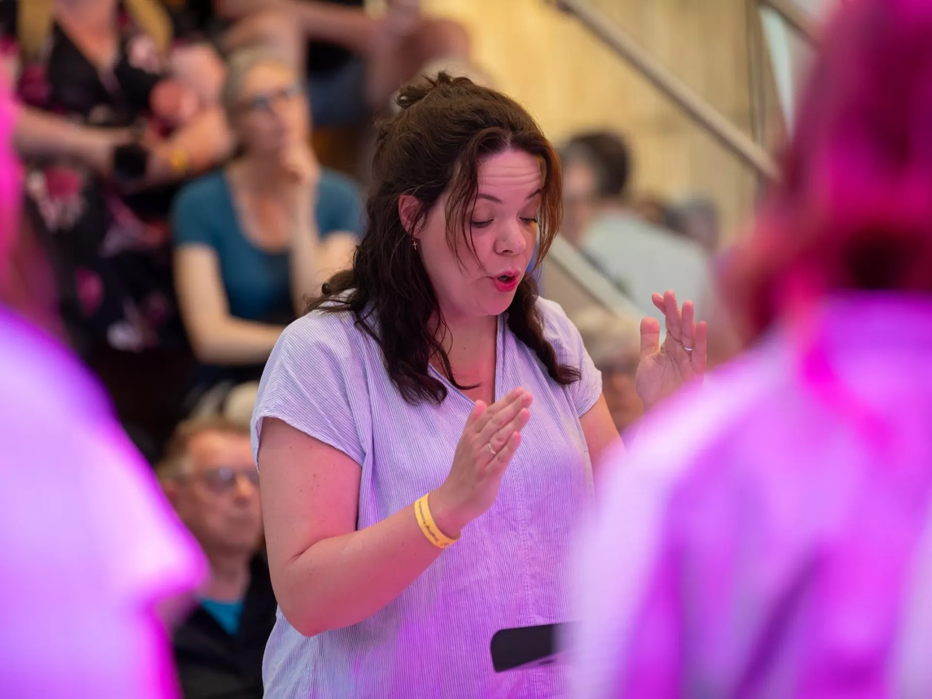Een vrouw dirigeert een groep tijdens een optreden, met een publiek op de achtergrond.