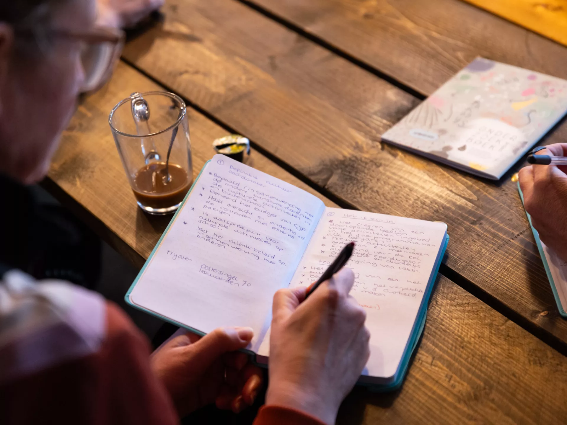 Persoon schrijft aantekeningen in een notitieboekje aan een houten tafel.