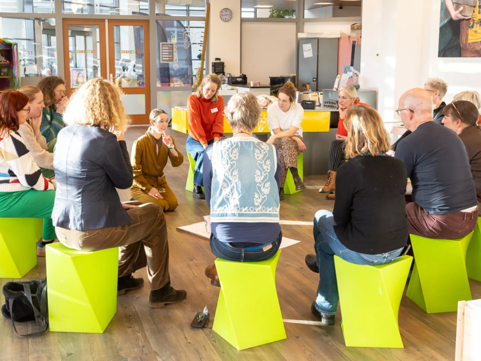 Een groep mensen zit in een kring op felgekleurde stoelen in een lichte, moderne ruimte.