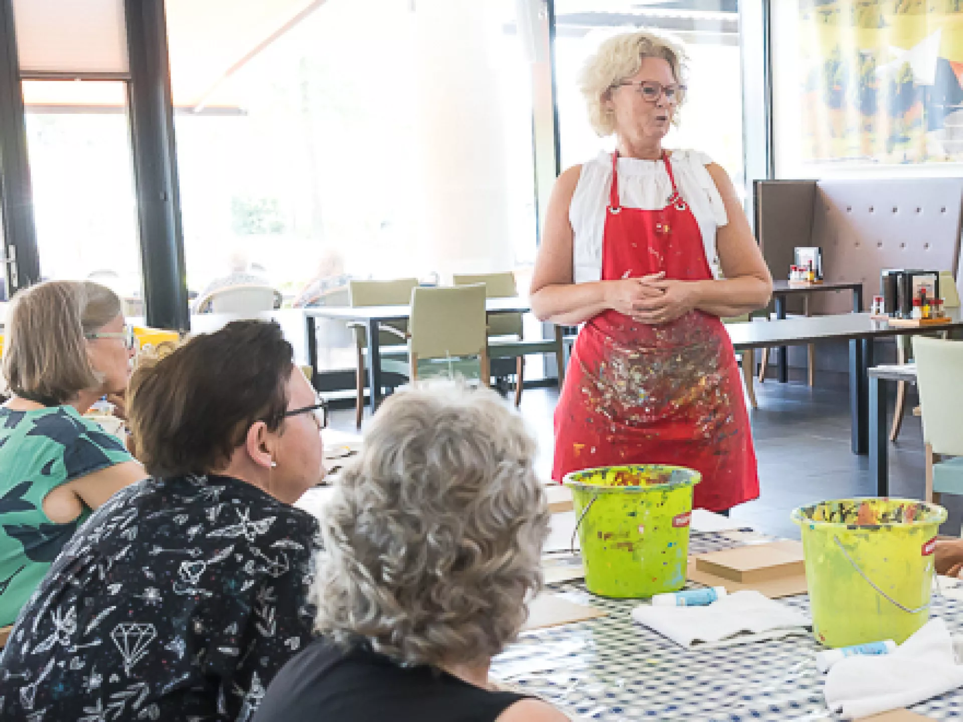 Verschillende mensen zitten aan een lange tafel. De docent beeldende kunst staat en geeft uitleg.