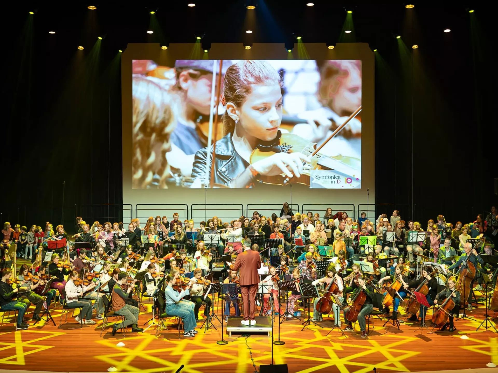 Symfonica in D. Een orkest voert een muziekvoorstelling uit op een fel verlicht podium met kinderen.