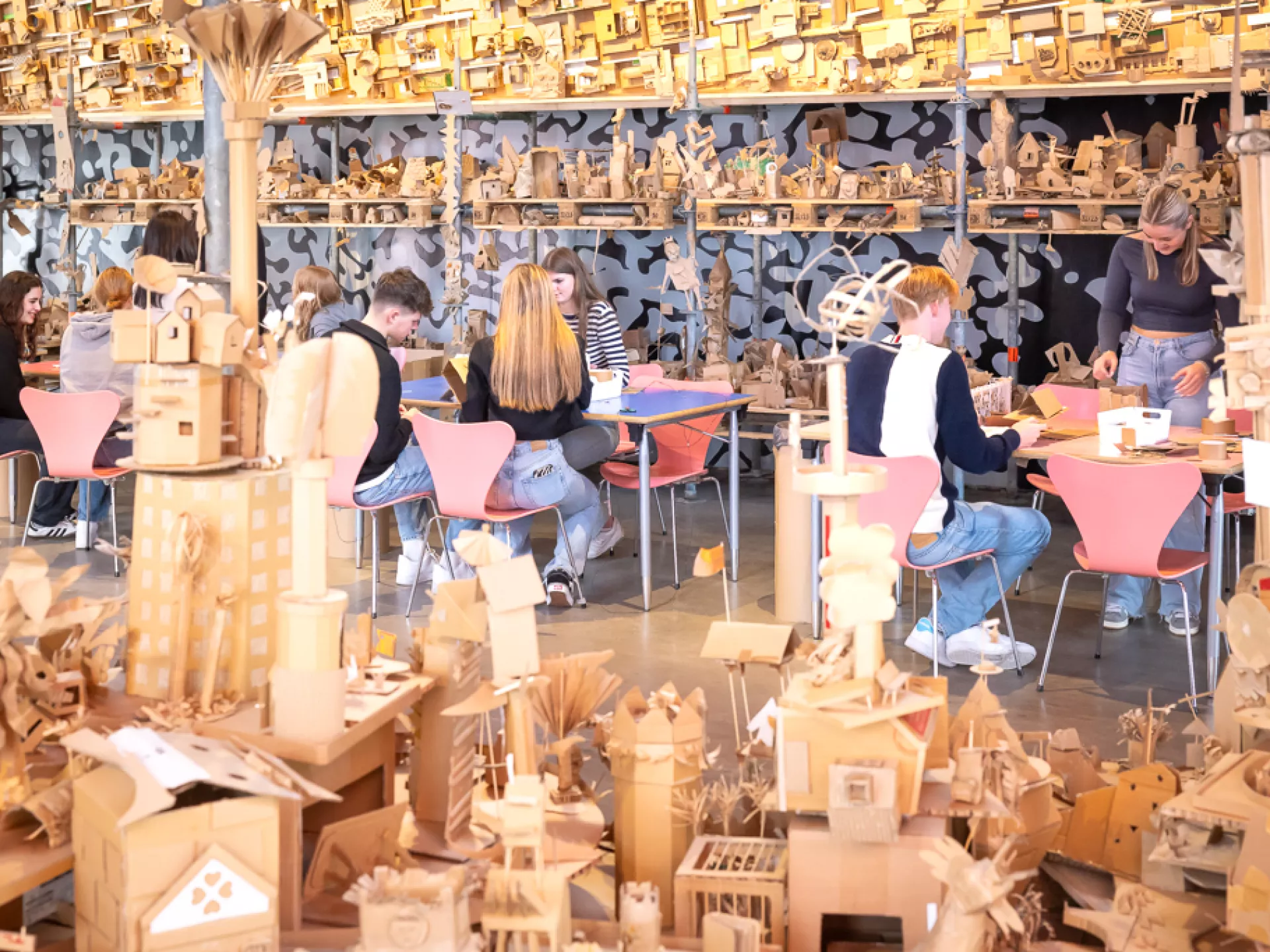 Mensen werken aan tafels, omringd door kartonnen kunstwerken in een creatieve ruimte.