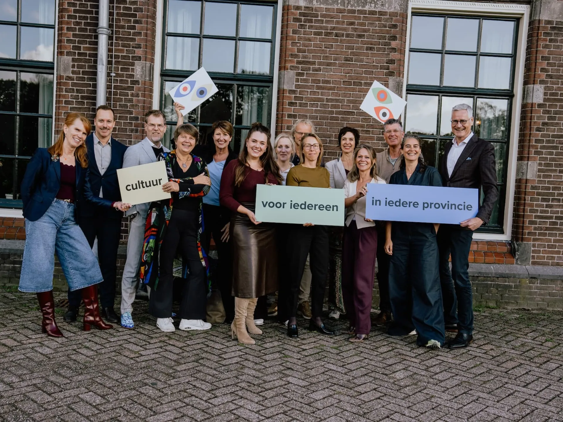 Een groep mensen poseert voor een gebouw, met borden en vlaggen.