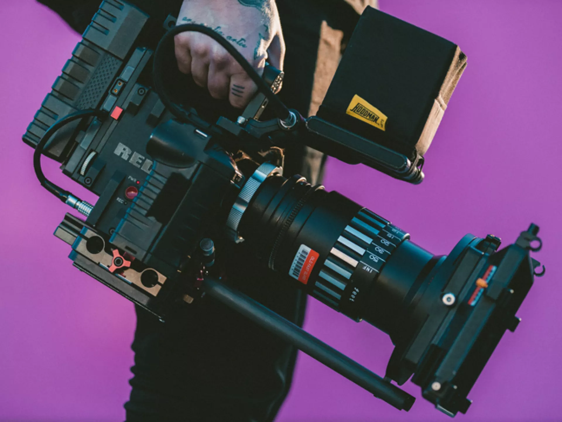 Een persoon houdt een professionele filmcamera vast tegen een paarse achtergrond.