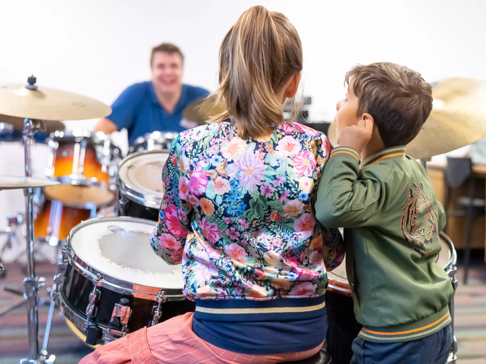 We zien twee kinderen op de rug zitten achter een drumstel met de docent ervoor. 