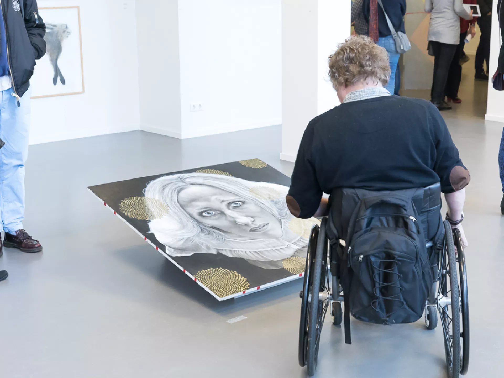 Een persoon in een rolstoel bekijkt een kunstwerk op de grond in een galerie.