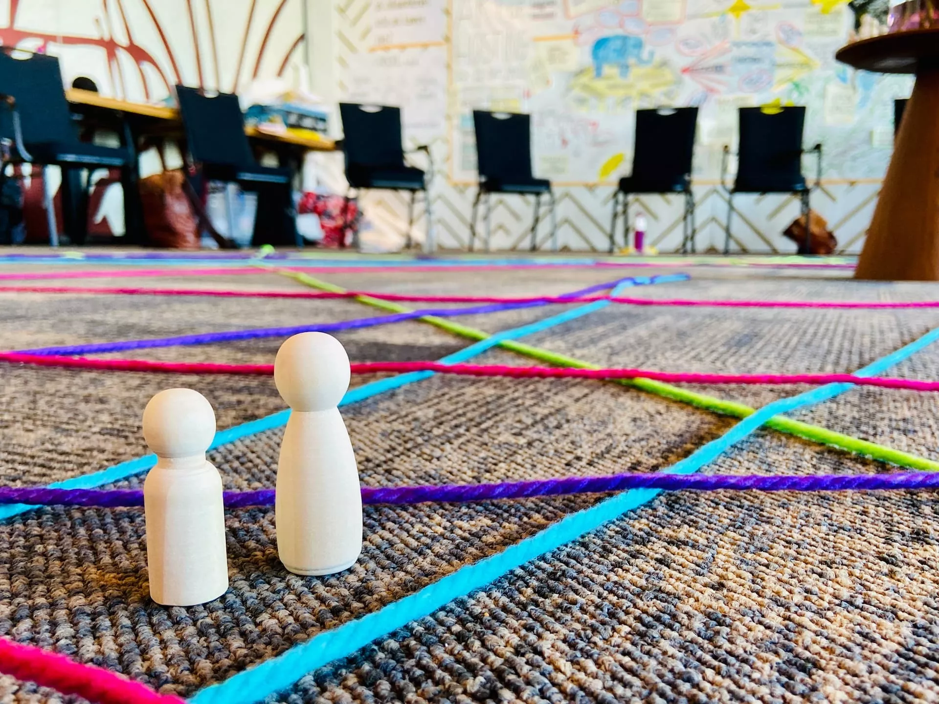 Twee houten pionnen staan op een vloer bedekt met kleurrijke touwpatronen, met op de achtergrond stoelen en een getekende muur.
