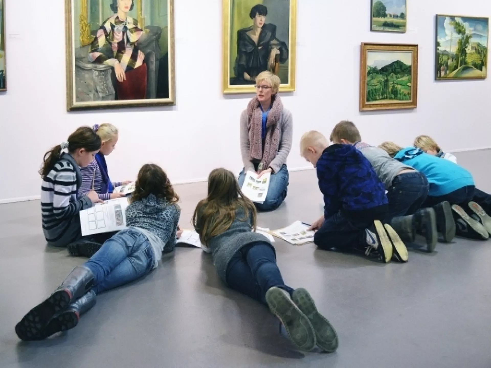Kinderen liggen en zitten in een halve cirkel om de docent op de grond in een museum. 
