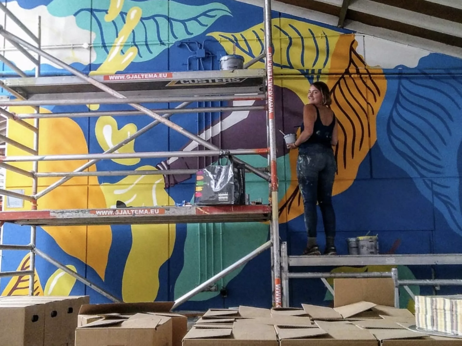 Kunstenaar schildert een kleurrijke muurschildering op een steiger in een magazijn.