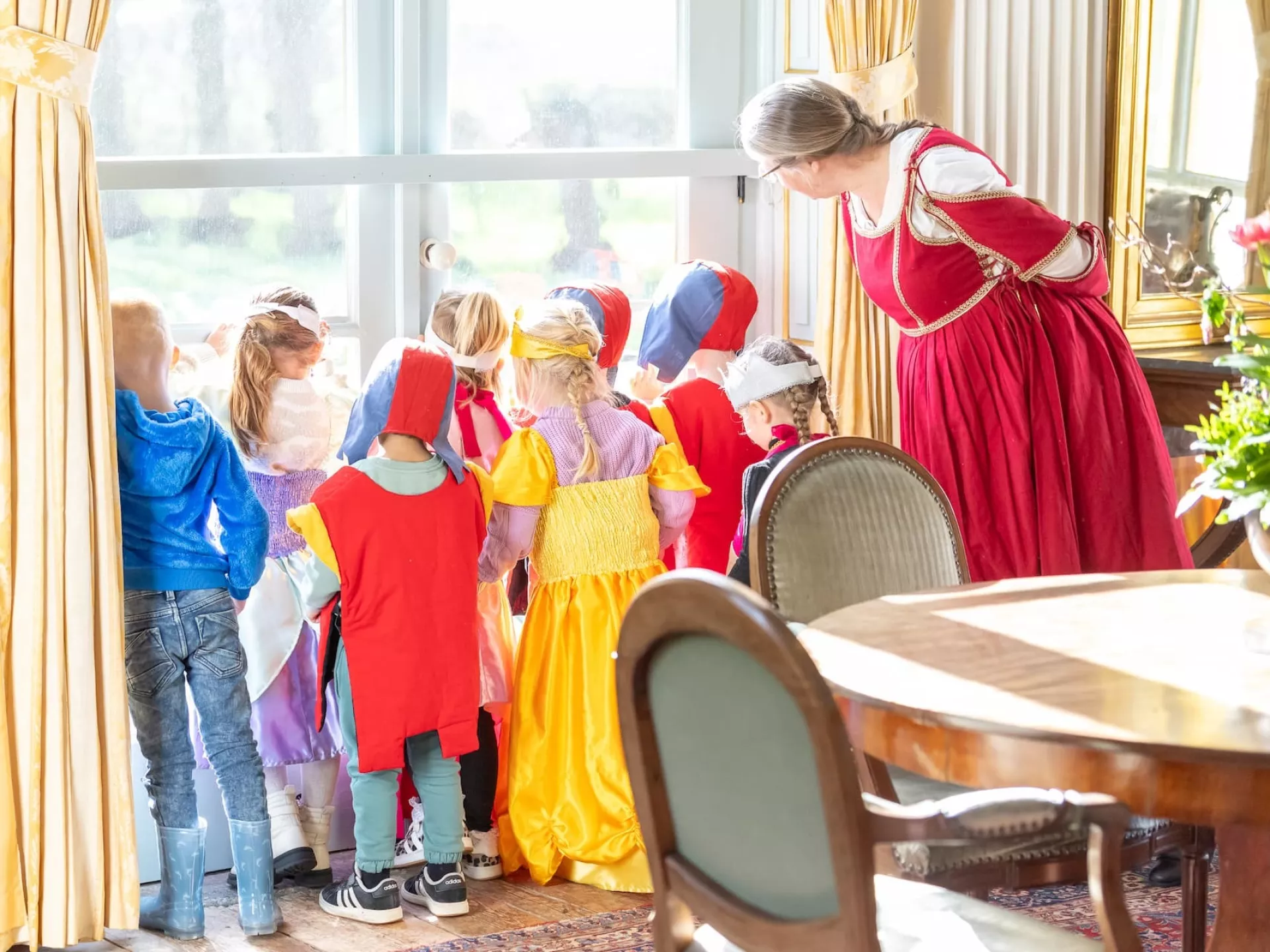 Kinderen en een volwassene in historische kostuums kijken nieuwsgierig uit het raam.