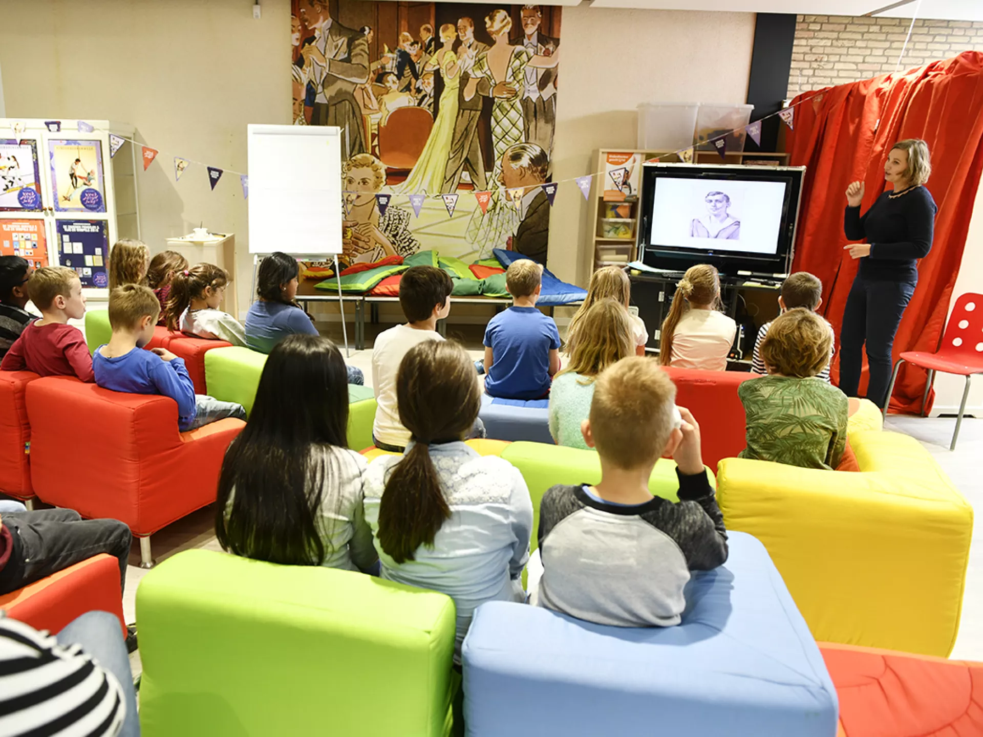 Groep kinderen op kleurrijke banken, luistert naar schrijfster