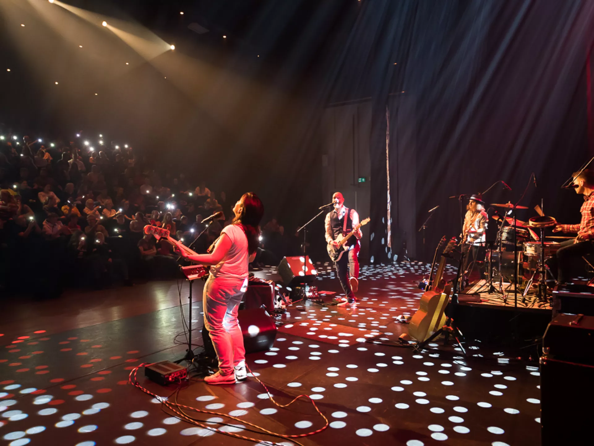 Een band treedt op in een concertzaal, met publiek dat hun mobiele telefoons omhoog houdt.
