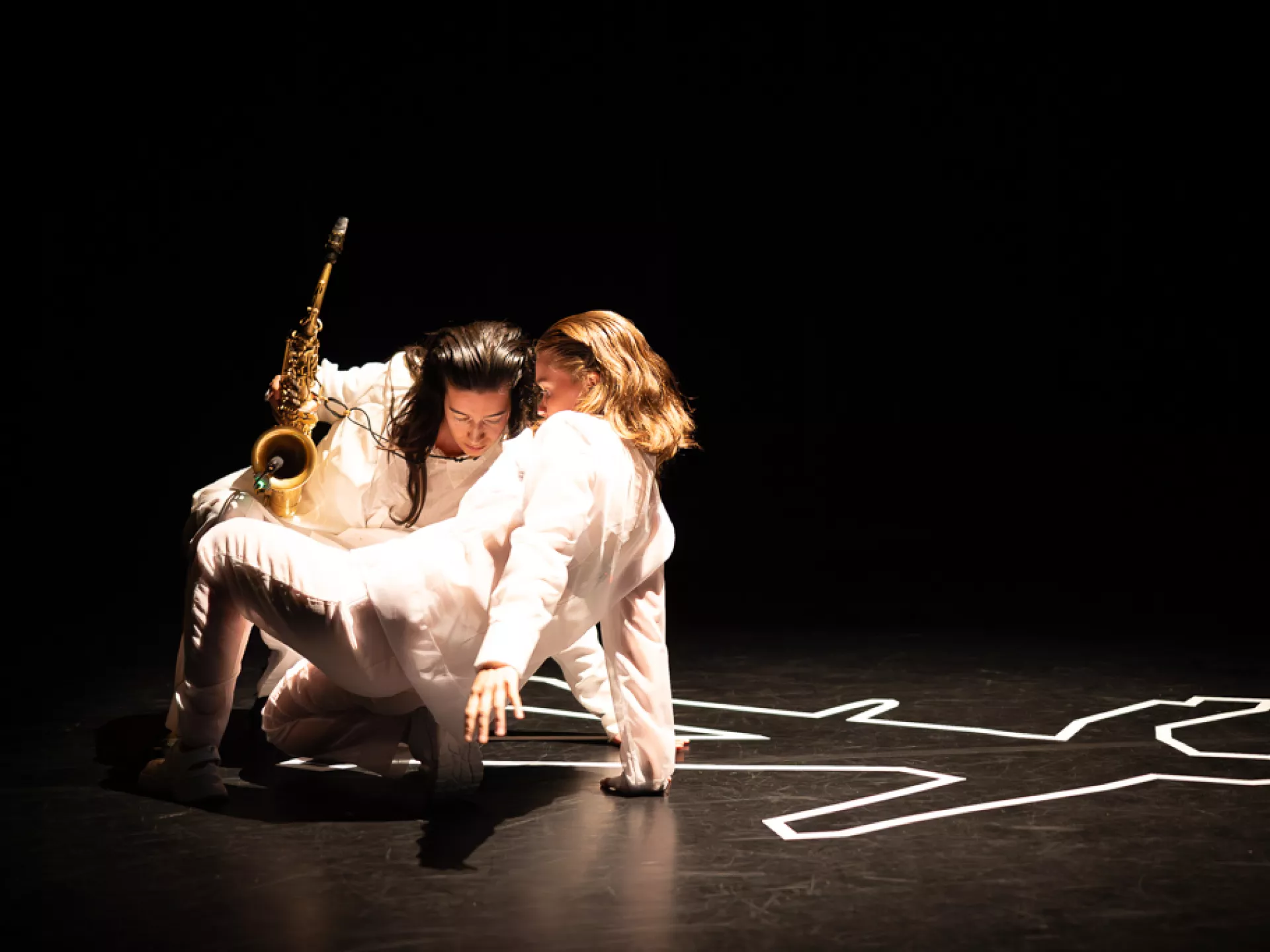 Twee performers op een donkere toneelvloer, waarbij een van hen een saxofoon vasthoudt naast een witte silhouet op de grond.