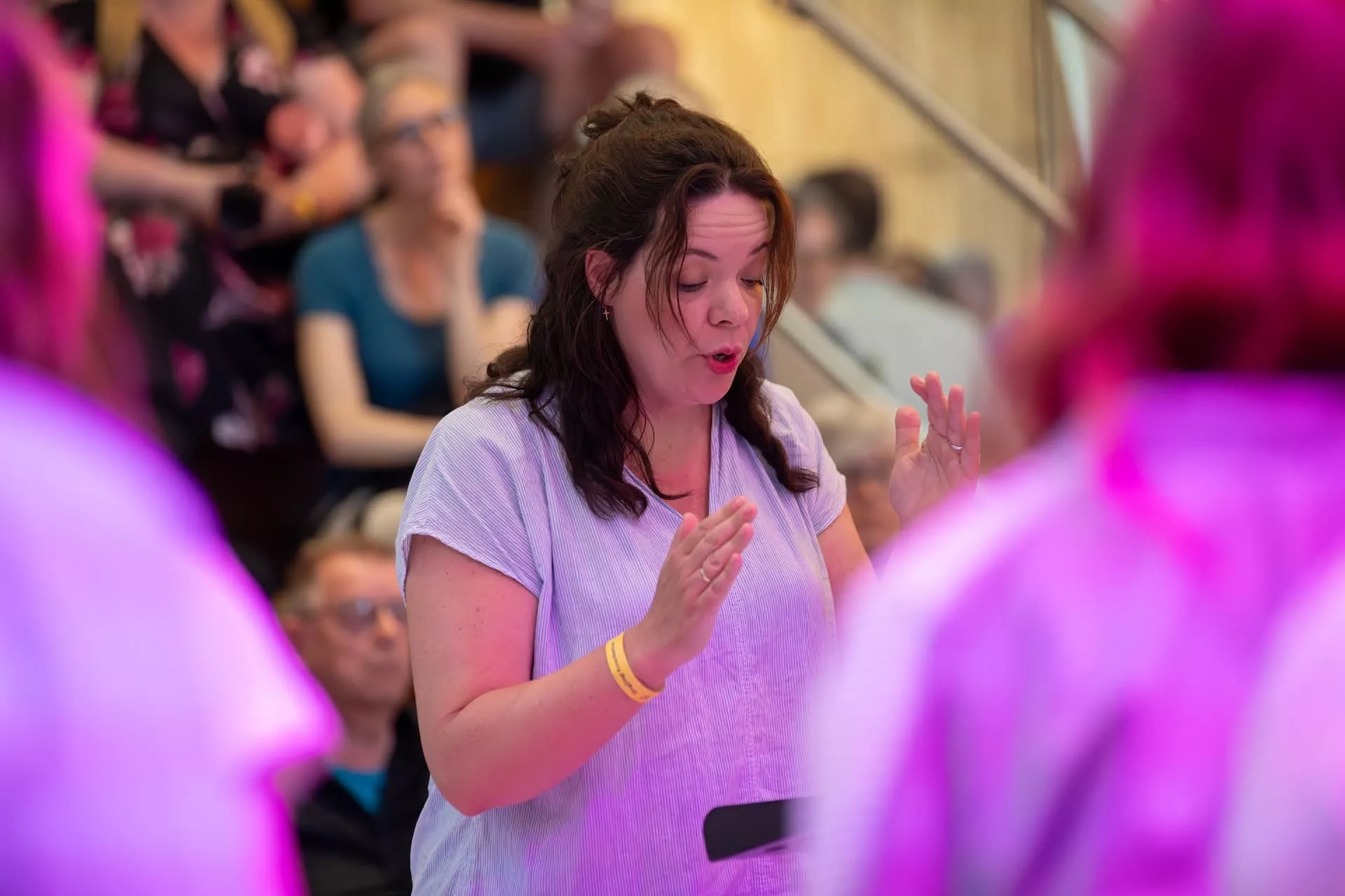 Een vrouw dirigeert een groep tijdens een optreden, met een publiek op de achtergrond.