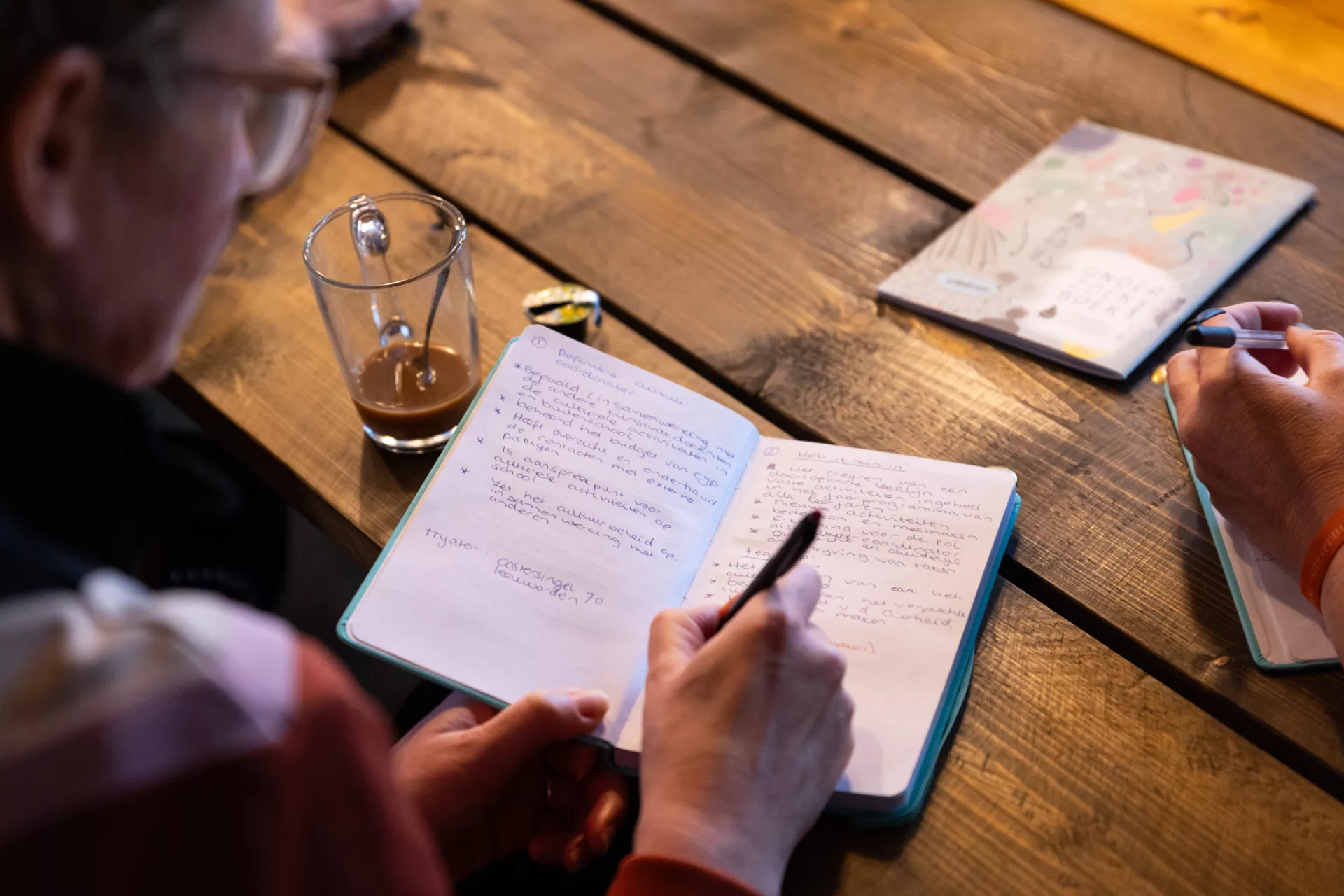 Persoon schrijft aantekeningen in een notitieboekje aan een houten tafel.