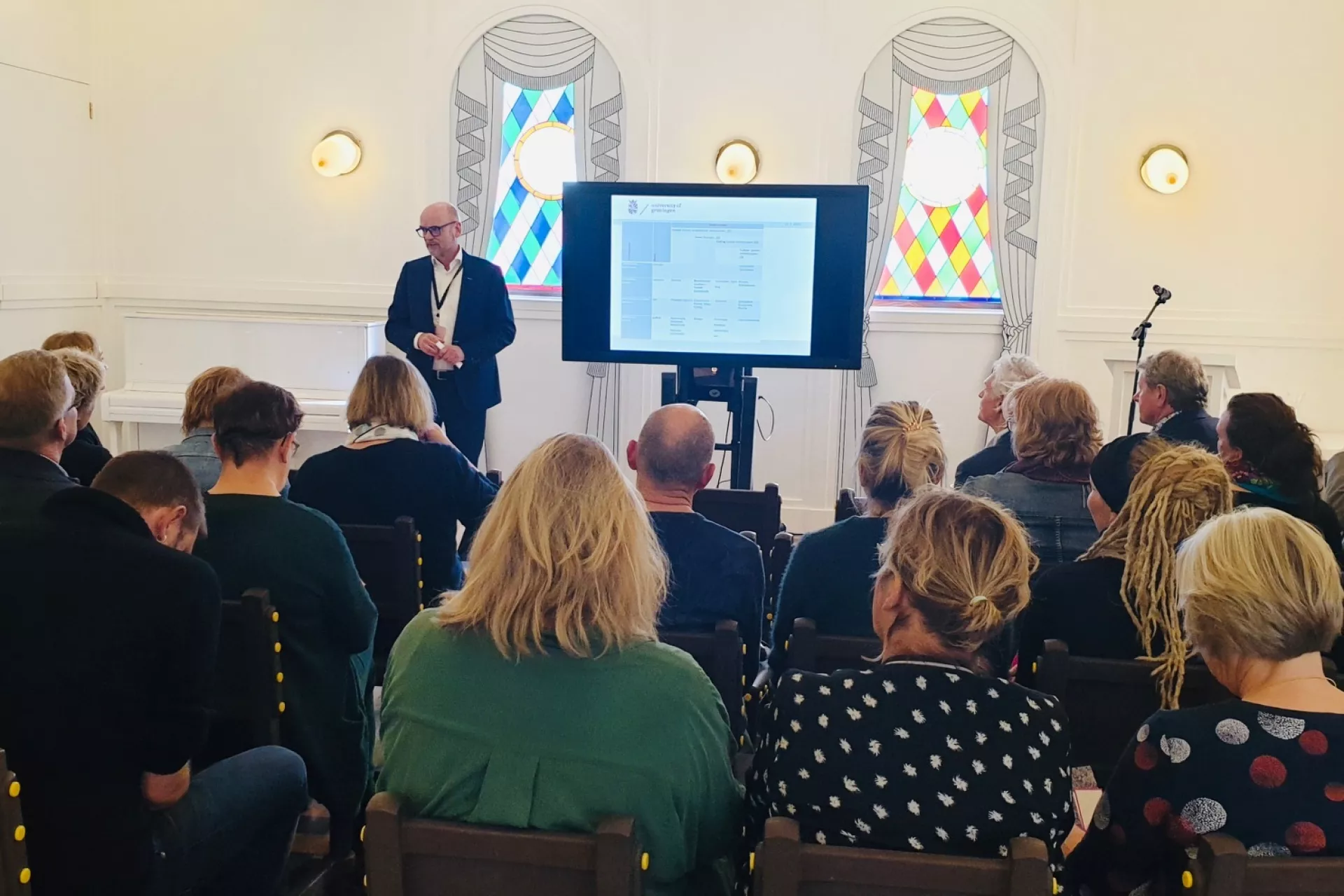 Een spreker geeft een presentatie aan een groep mensen in een zaal met glas-in-loodramen.