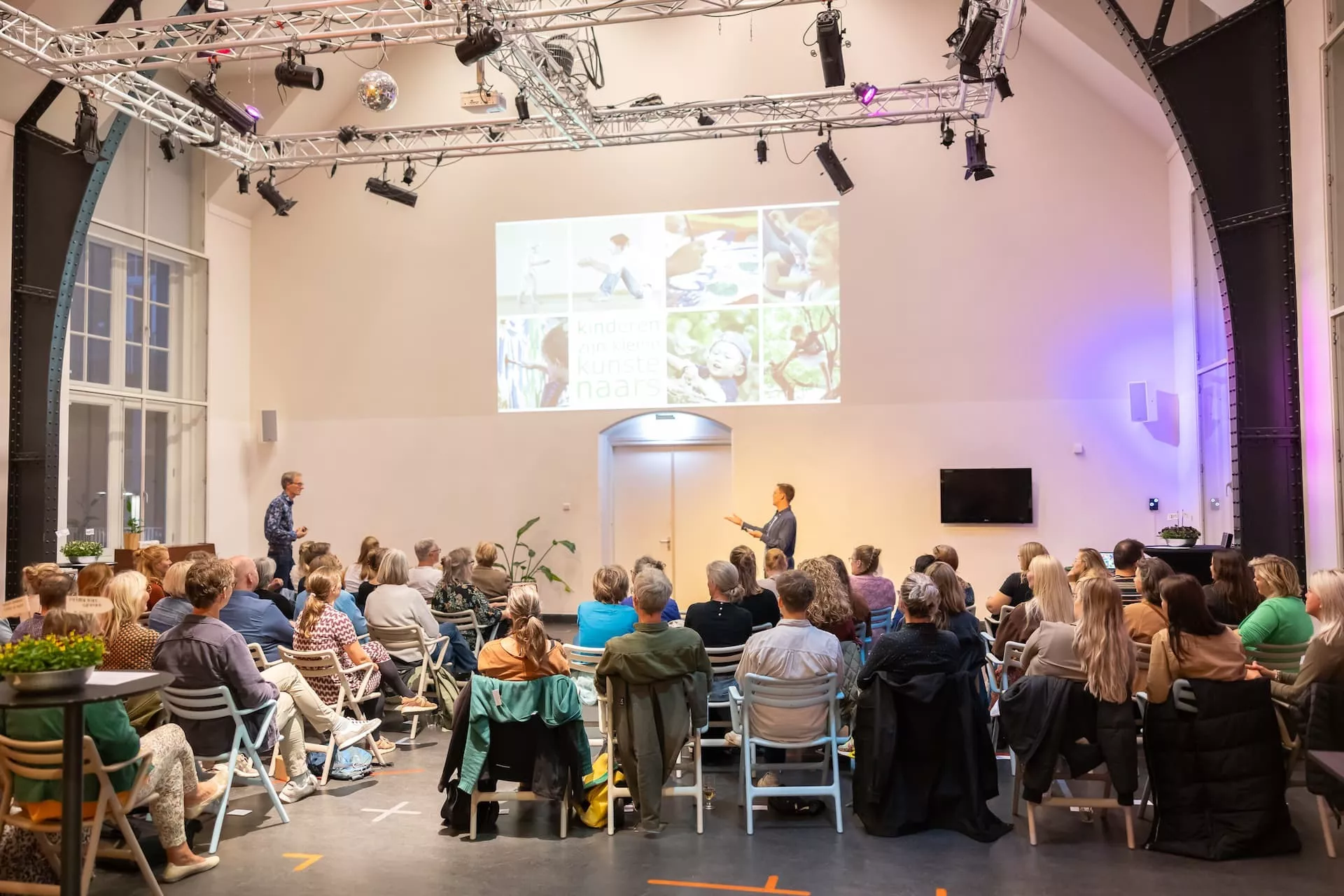 Mensen luisteren naar een presentatie in een verlichte zaal.
