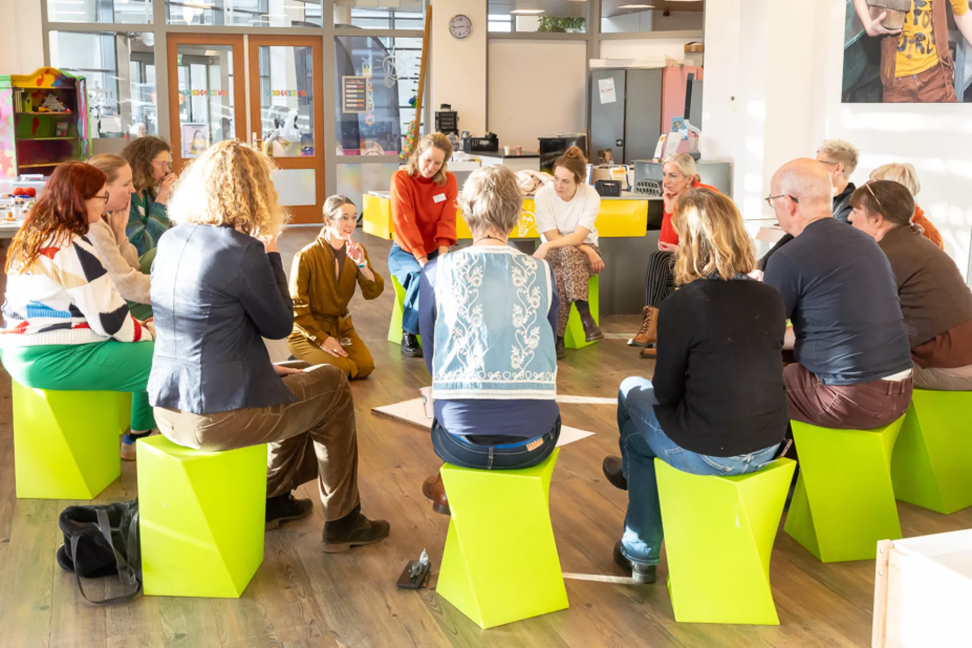 Een groep mensen zit in een kring op felgekleurde stoelen in een lichte, moderne ruimte.