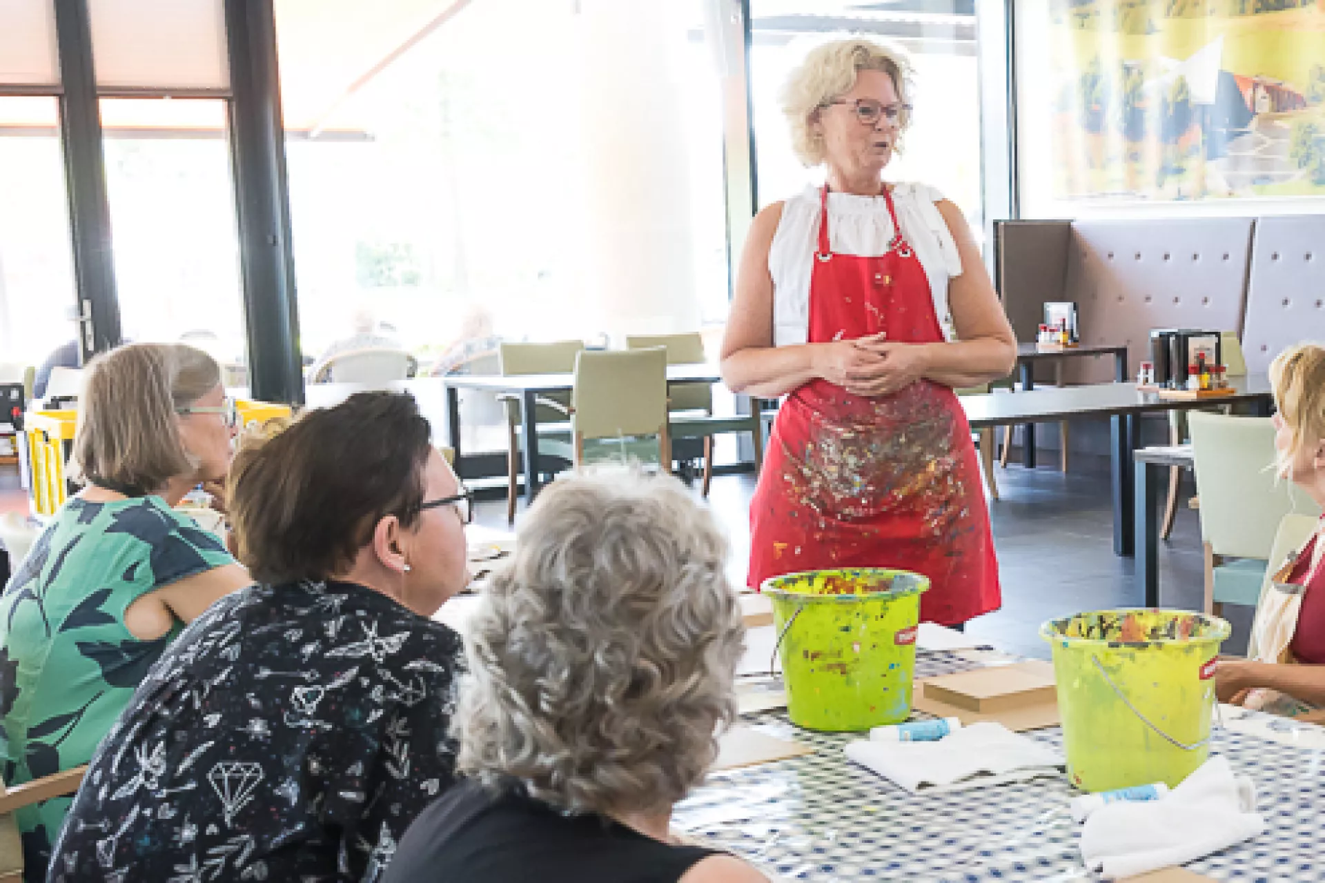 Verschillende mensen zitten aan een lange tafel. De docent beeldende kunst staat en geeft uitleg.