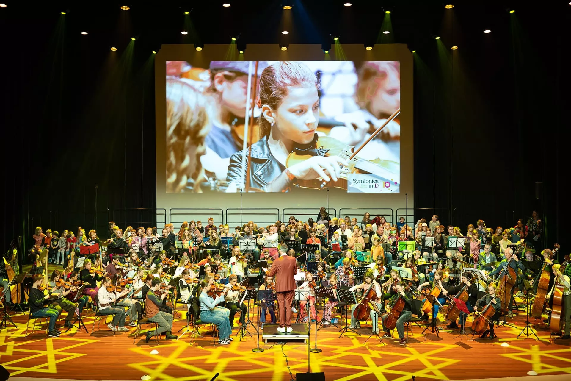 Symfonica in D. Een orkest voert een muziekvoorstelling uit op een fel verlicht podium met kinderen.