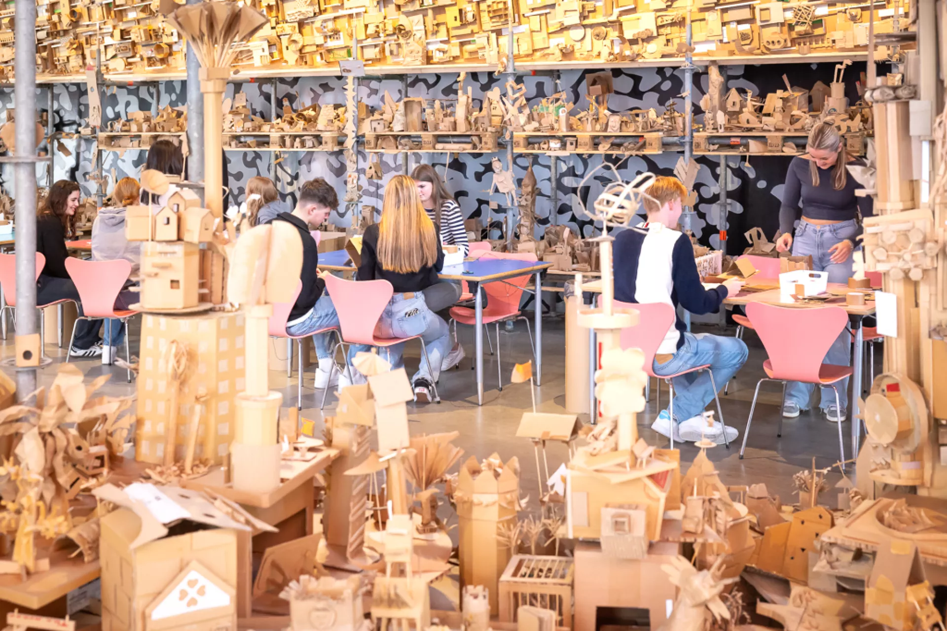 Mensen werken aan tafels, omringd door kartonnen kunstwerken in een creatieve ruimte.