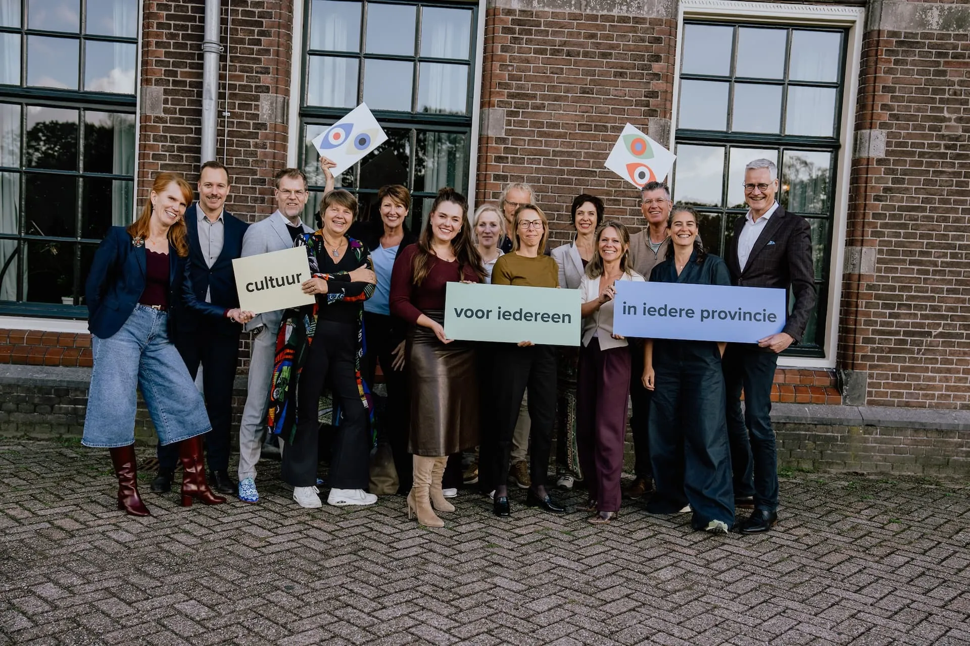 Een groep mensen poseert voor een gebouw, met borden en vlaggen.