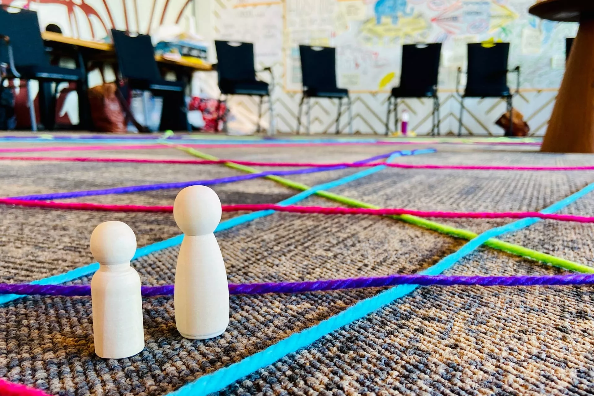 Twee houten pionnen staan op een vloer bedekt met kleurrijke touwpatronen, met op de achtergrond stoelen en een getekende muur.