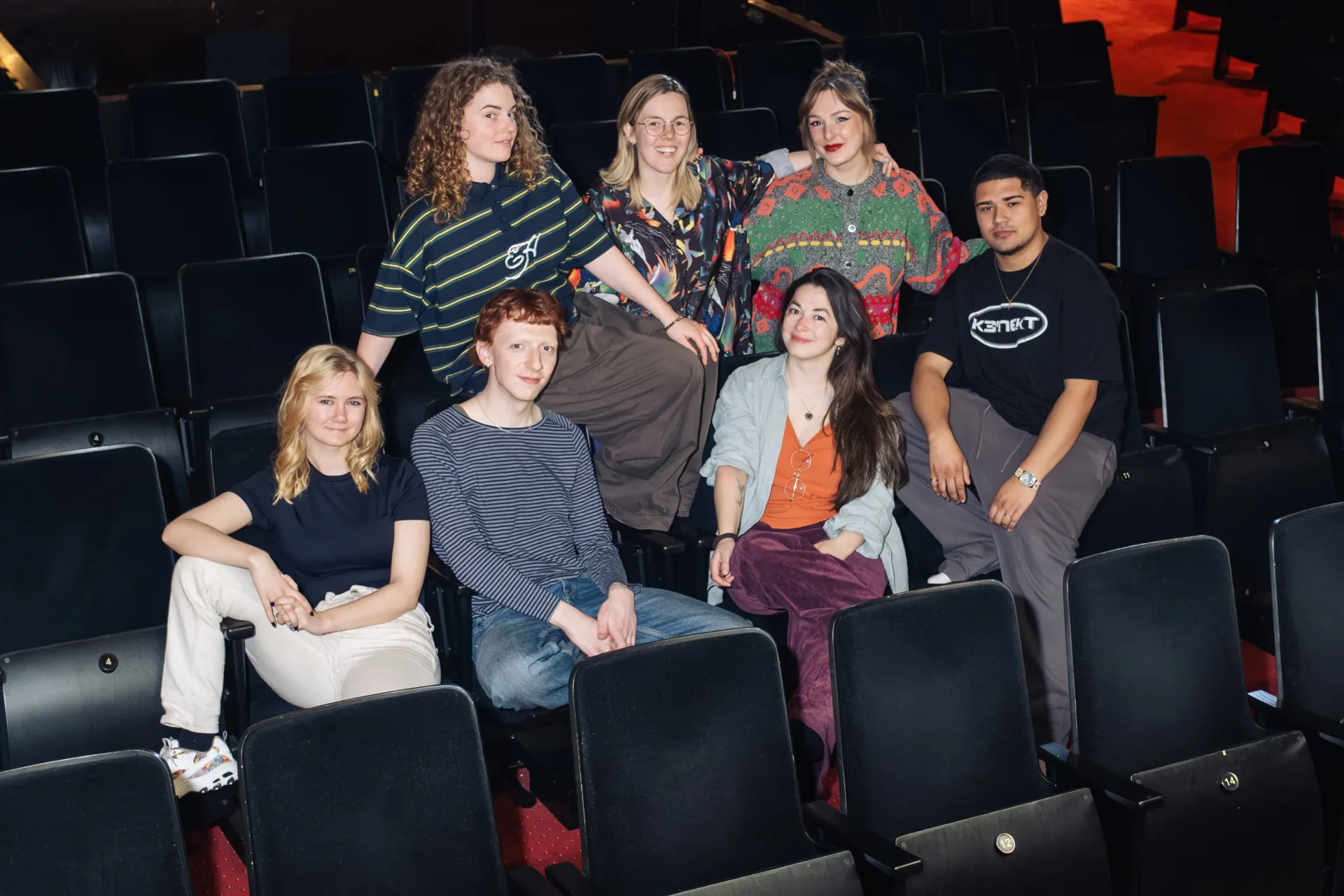 Een groep jonge mensen zit samen in een lege theaterzaal.