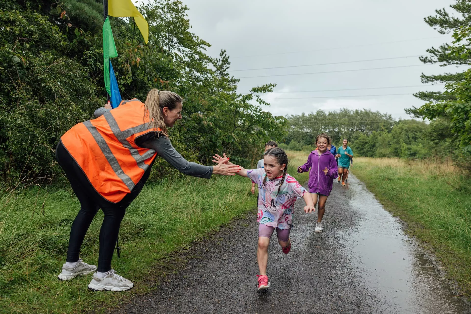 Kinderen rennen op een verharde weg en geven een high five aan een begeleider in de berm in een fluoriserend hesje