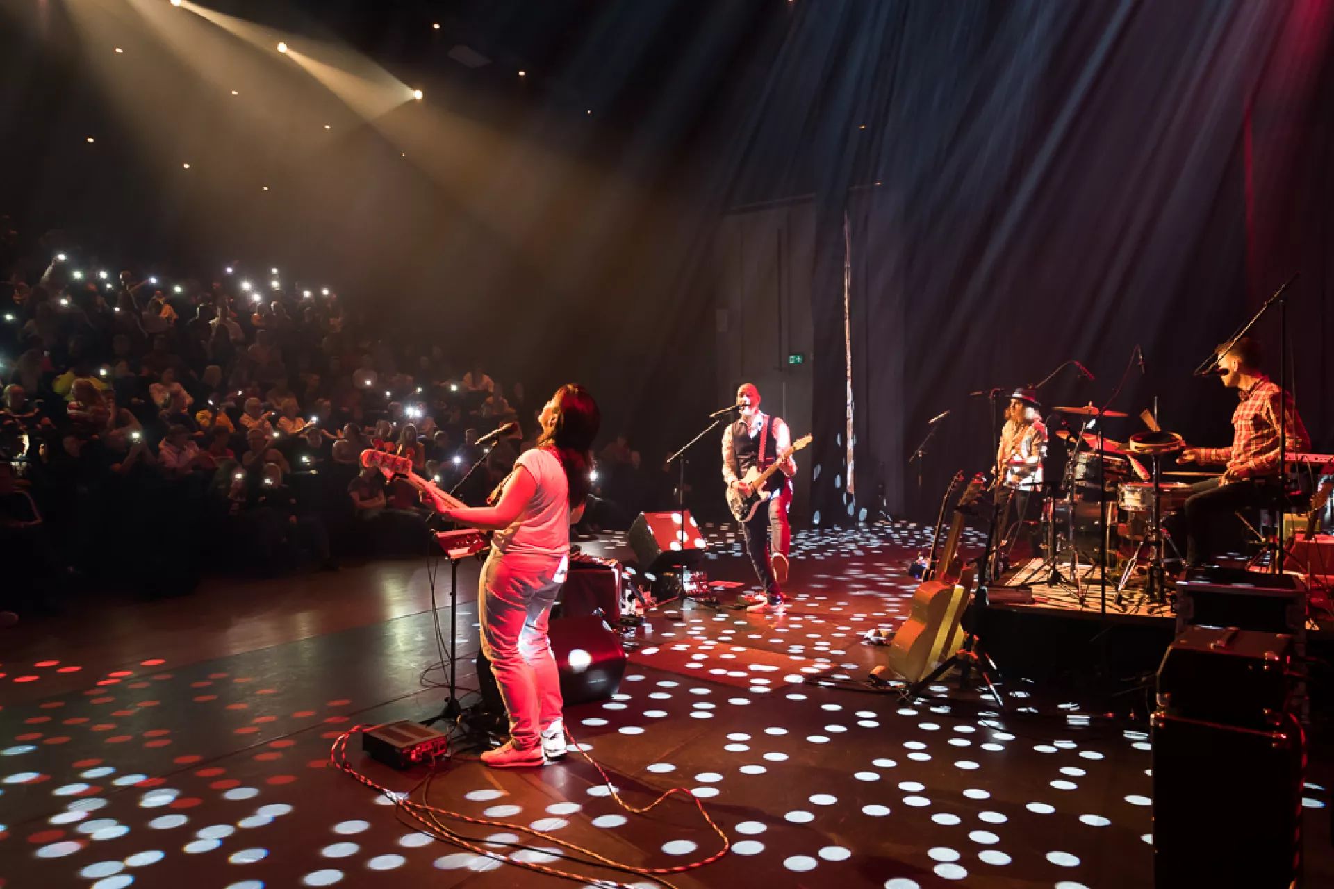 Een band treedt op in een concertzaal, met publiek dat hun mobiele telefoons omhoog houdt.