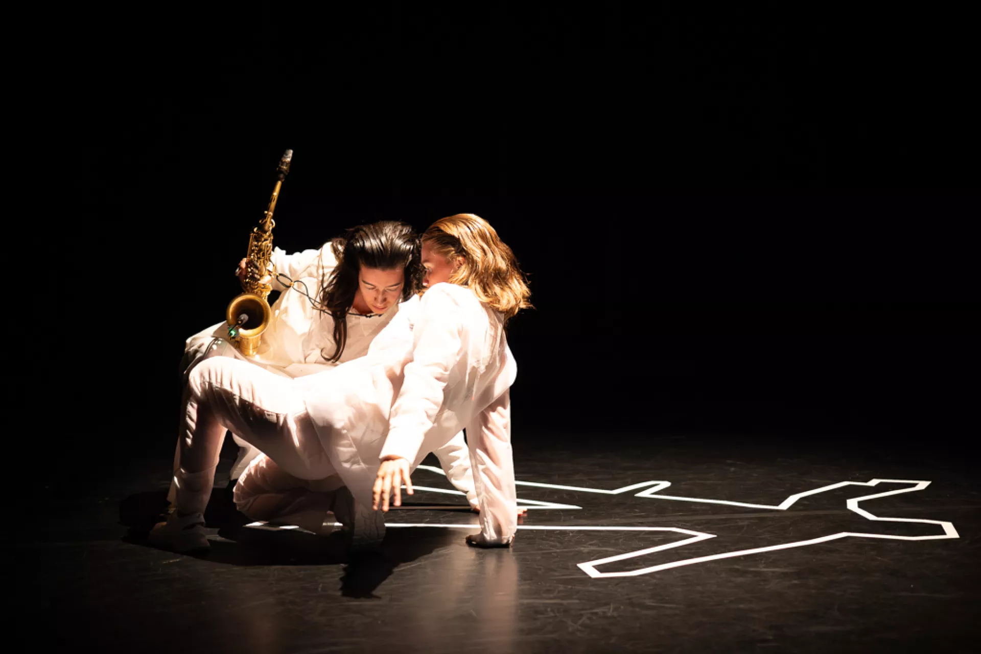 Twee performers op een donkere toneelvloer, waarbij een van hen een saxofoon vasthoudt naast een witte silhouet op de grond.